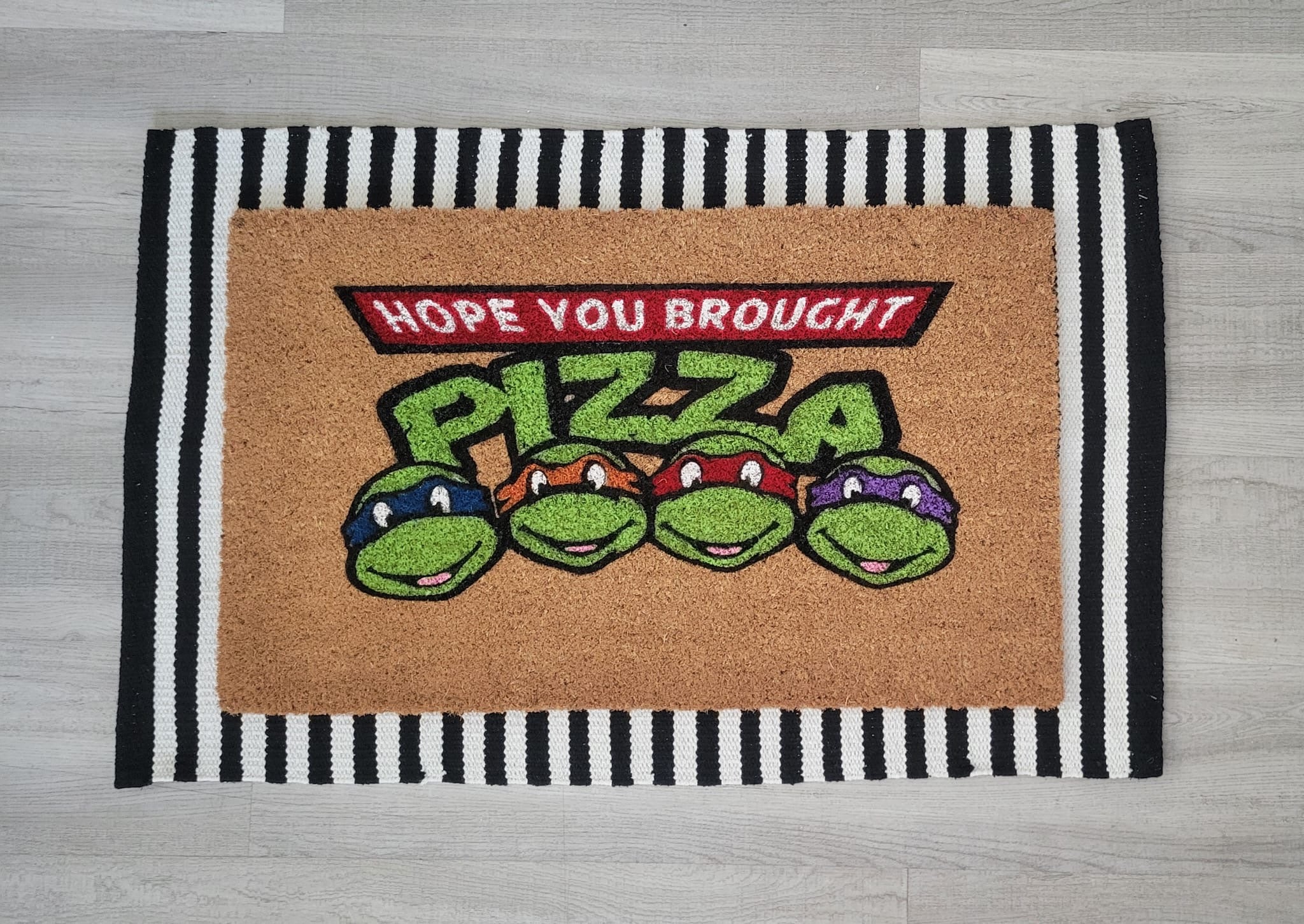 Coir doormat with the phrase ‘Hope You Brought Pizza,’ hand-painted in bold, playful lettering with a Ninja Turtle–inspired design